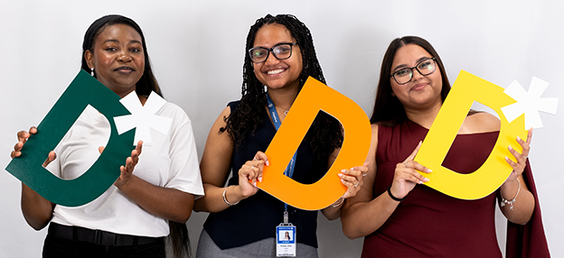 Three female students hold up the letter D in front of a white background smiling.